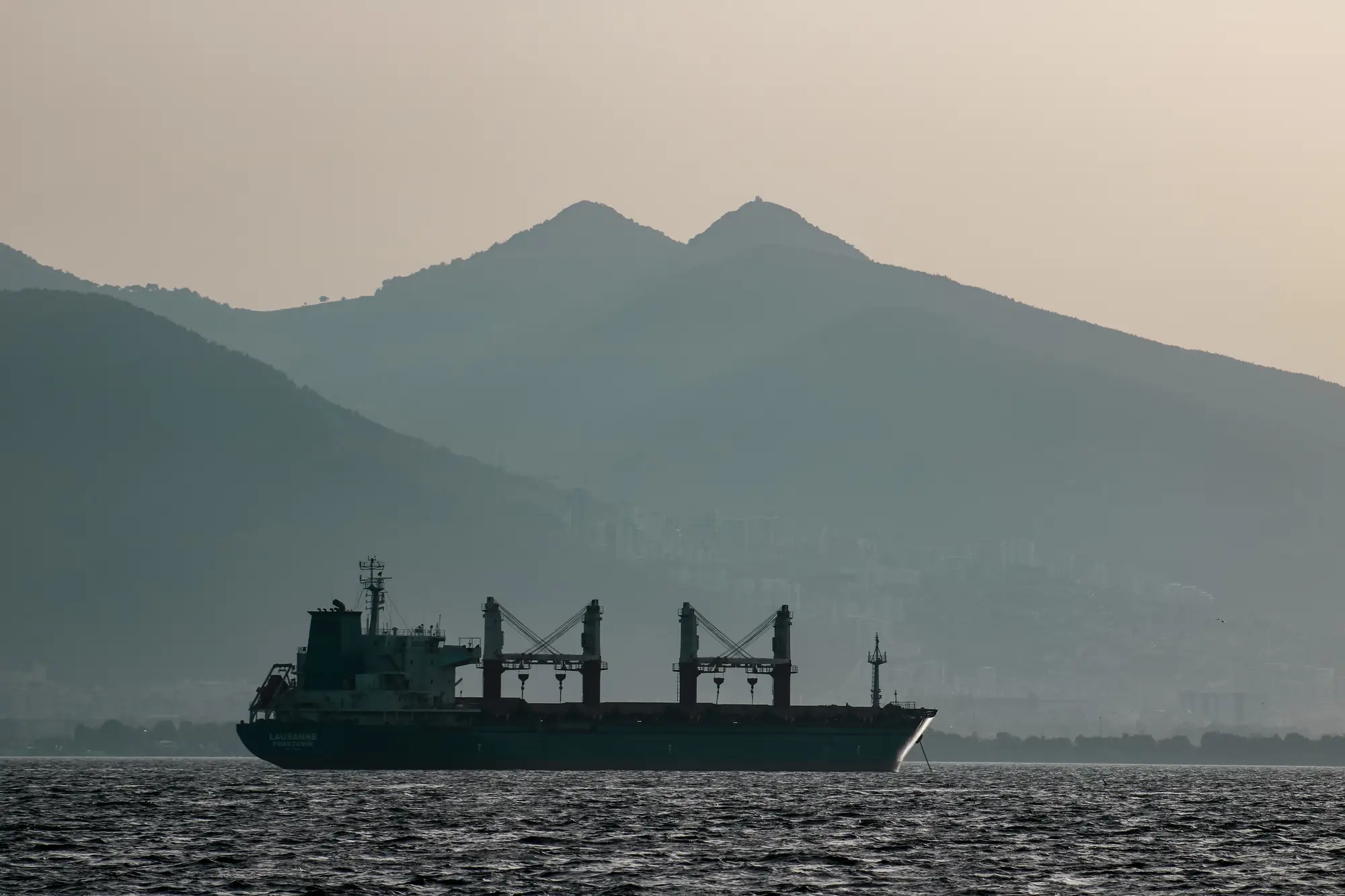 A geared bulk carrier at anchor in a hazy bay, mountains behind it