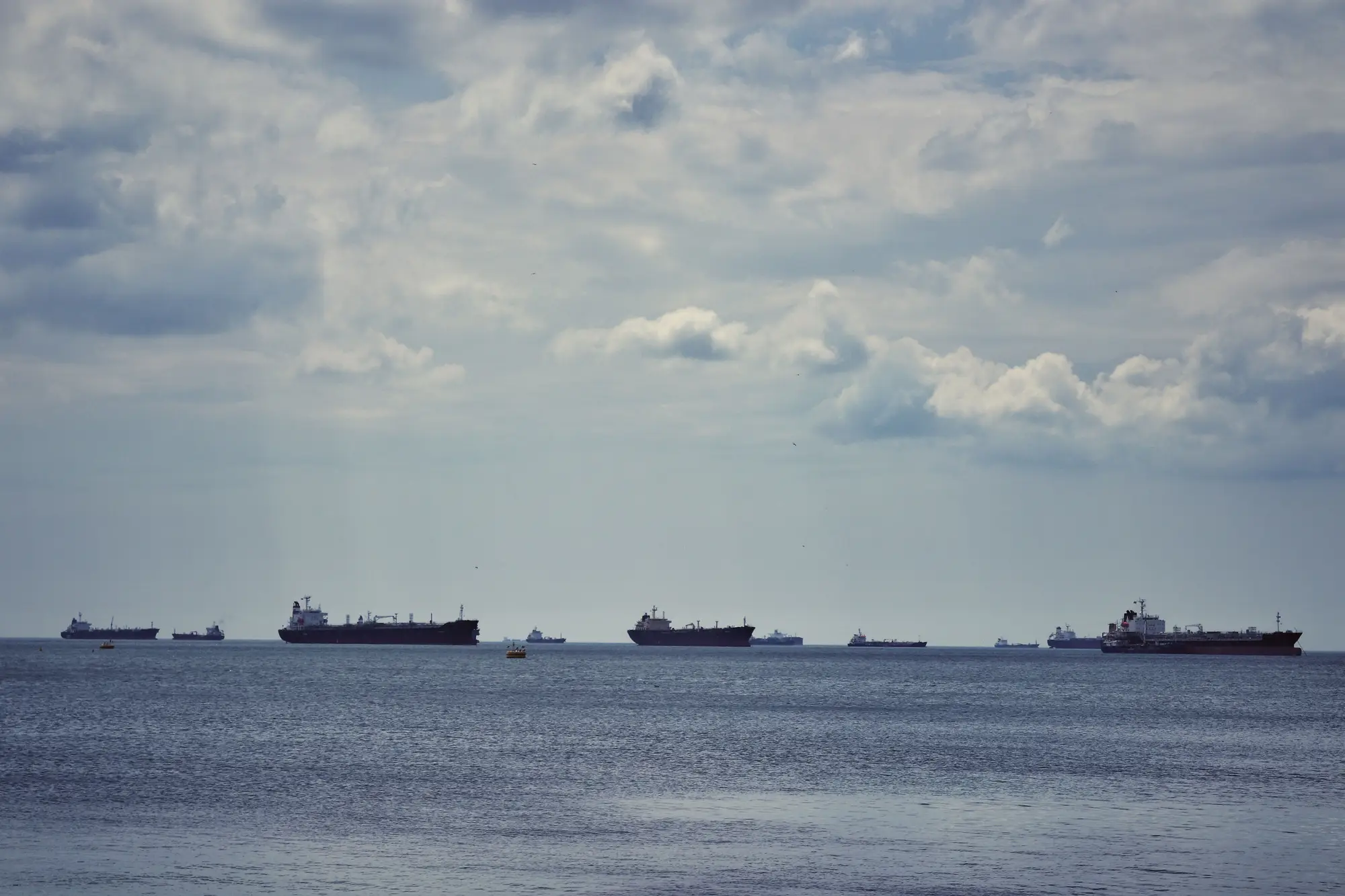 A line of cargo ships and tankers at anchor offshore, spread along the horizon under a grey, broken sky